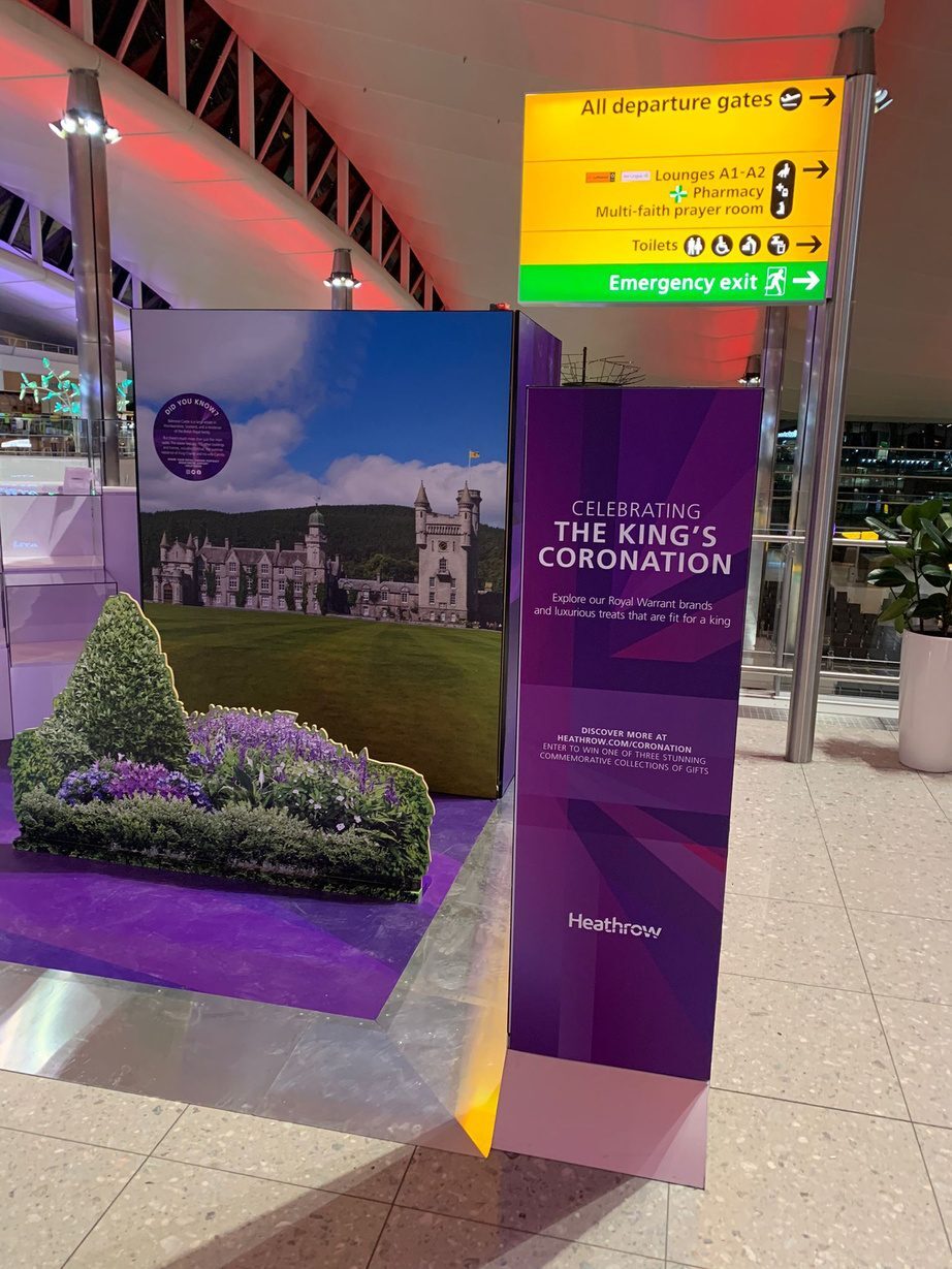 Heathrow King's Coronation display totem beside a Balmoral Castle backdrop and floral planting, under departure gate signage